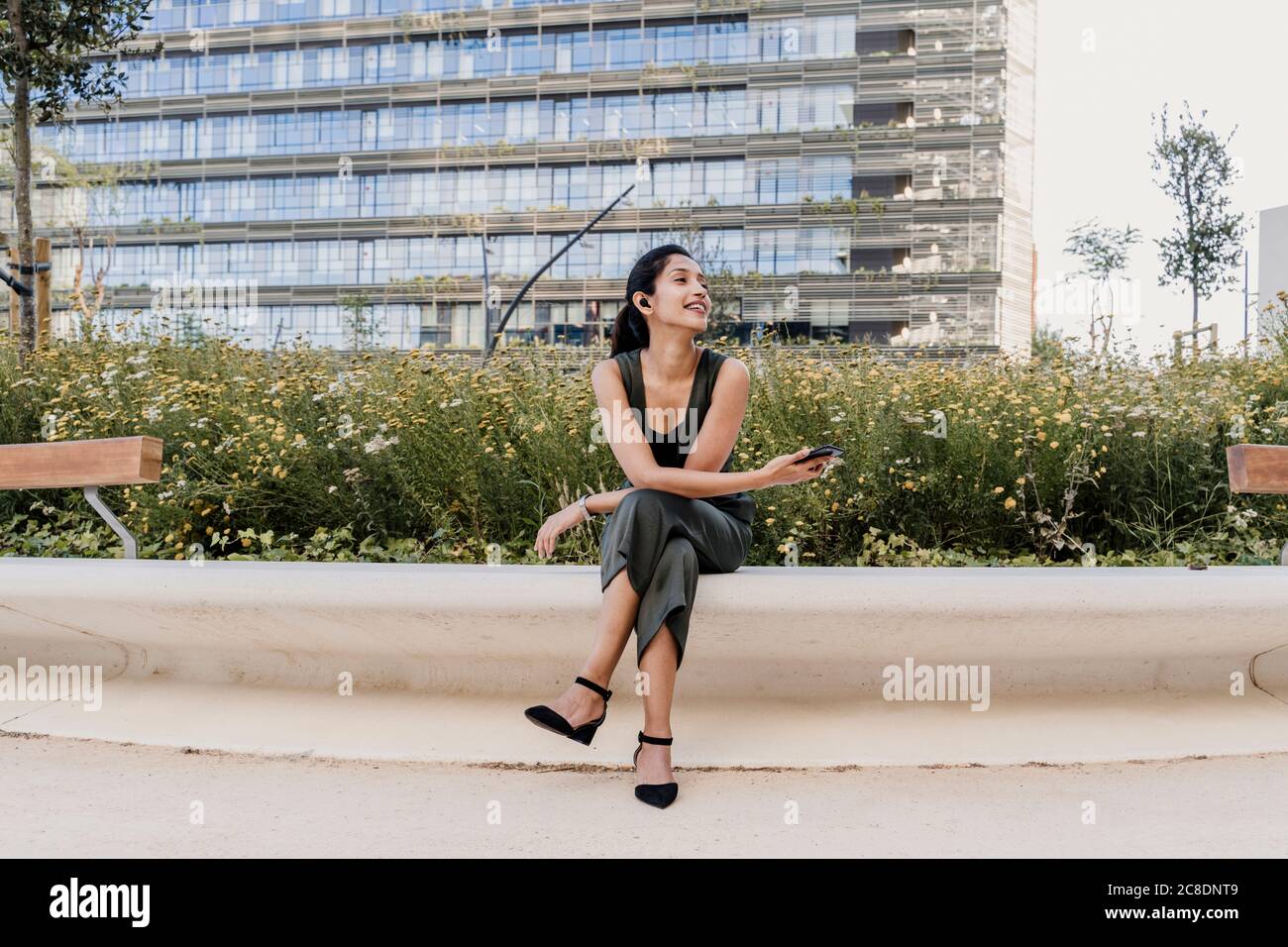 Lächelnde Geschäftsfrau hält Smartphone und schaut weg, während sie sitzt Sitz gegen Pflanzen Stockfoto