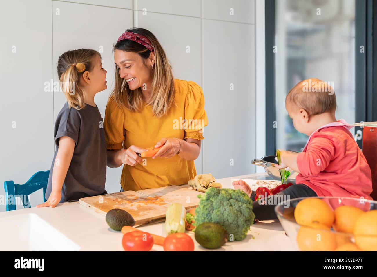 Lächelnde Mutter und Mädchen, die Nahrung während Baby Tochter spielen Auf Kücheninsel Stockfoto