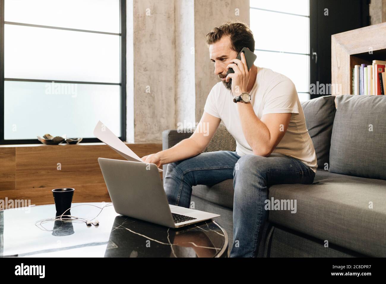 Reifer Mann auf der Couch sitzend, mit Laptop, am Telefon sprechend, Papiere haltend Stockfoto