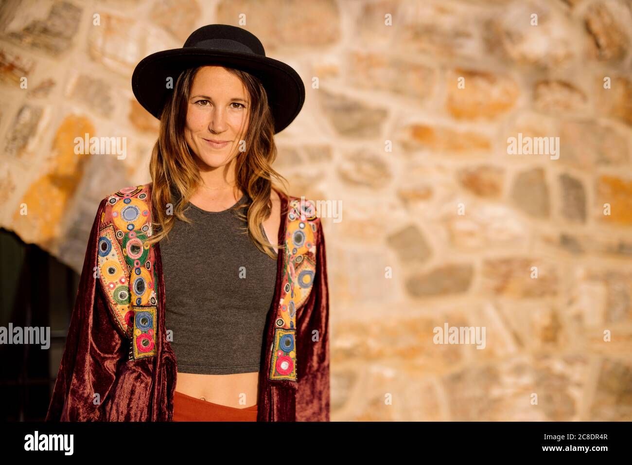 Selbstbewusste Frau trägt Hut und Jacke stehen gegen Steinwand An sonnigen Tagen Stockfoto