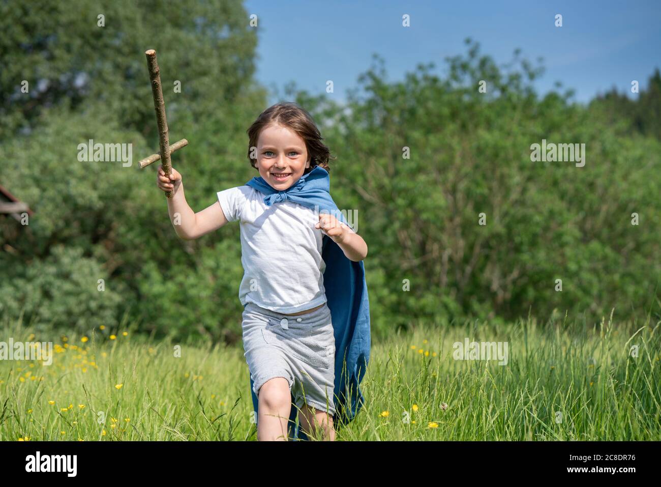 Verspielter Junge trägt Umhang mit Spielzeug Schwert beim Laufen auf Grasland Stockfoto