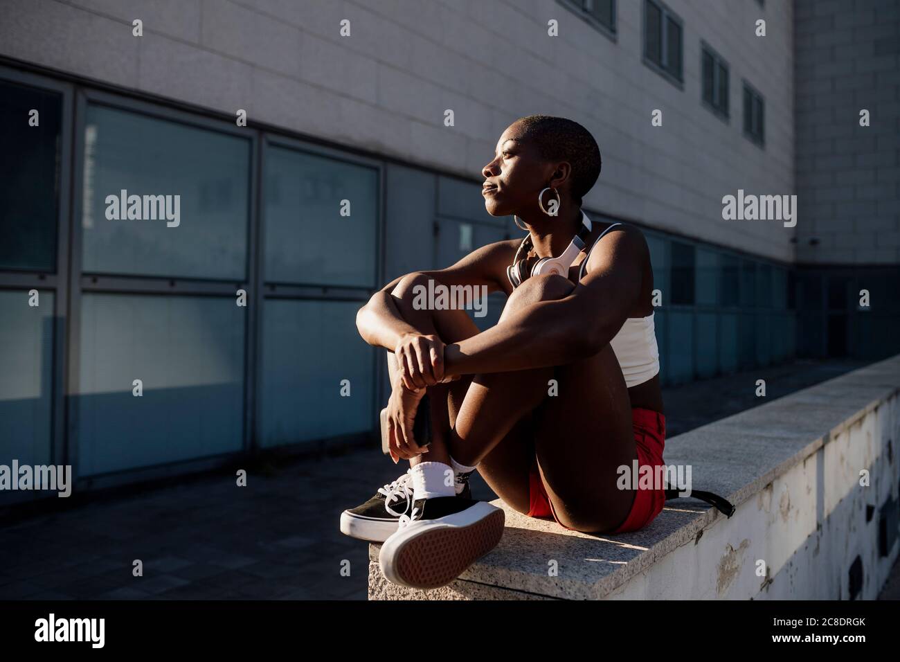 Nachdenkliche junge Frau mit rasierten Kopf sitzt auf Stützwand Gegen Gebäude Stockfoto