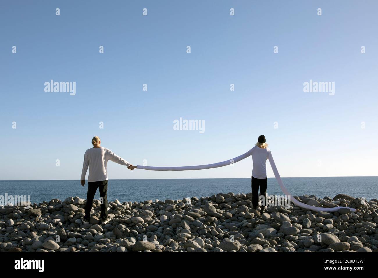 Frau mit langen künstlichen Armen hält Mann die Hand auf Kieselsteinen Am Strand gegen klaren Himmel Stockfoto