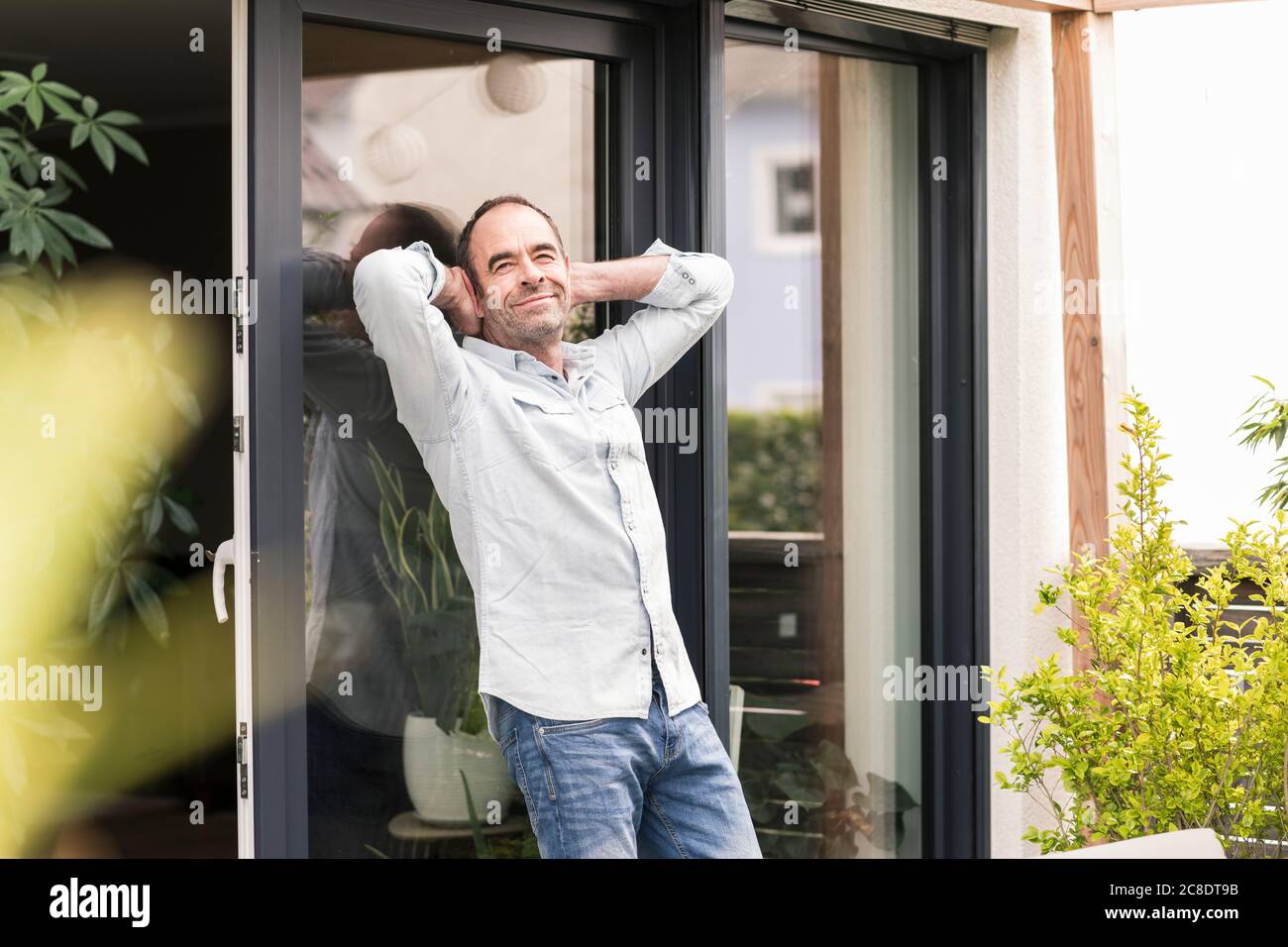 Lächelnder reifer Mann, der sich am Fenster mit den Händen hinter dem Kopf lehnte Im Hinterhof Stockfoto