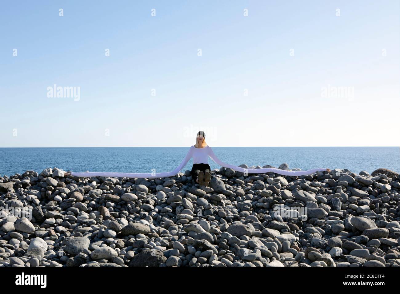 Frau mit künstlichen langen Händen sitzt auf Kieselsteinen am Strand Gegen klaren Himmel Stockfoto