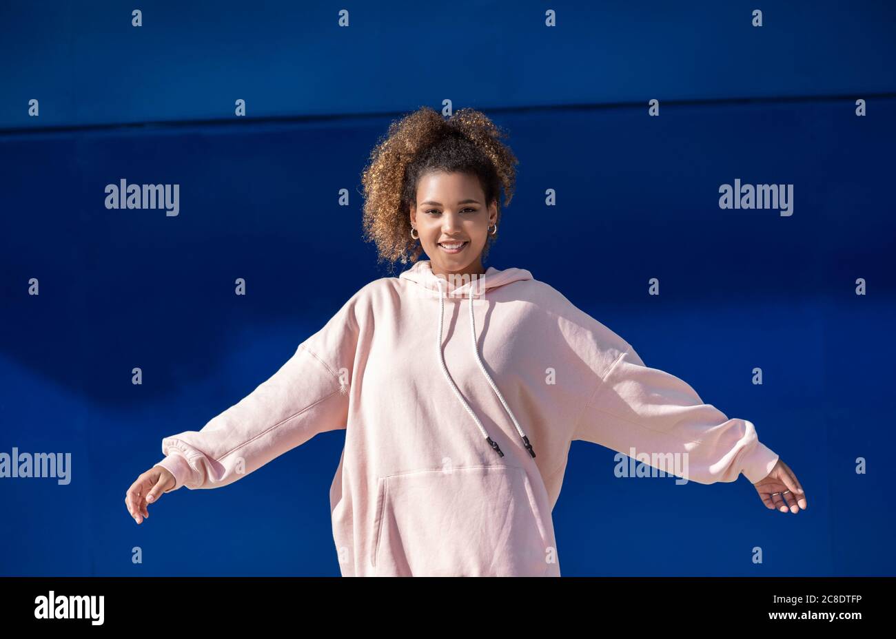 Lächelnde junge Frau trägt rosa Kapuzenpullover vor Blaue Wand Stockfoto