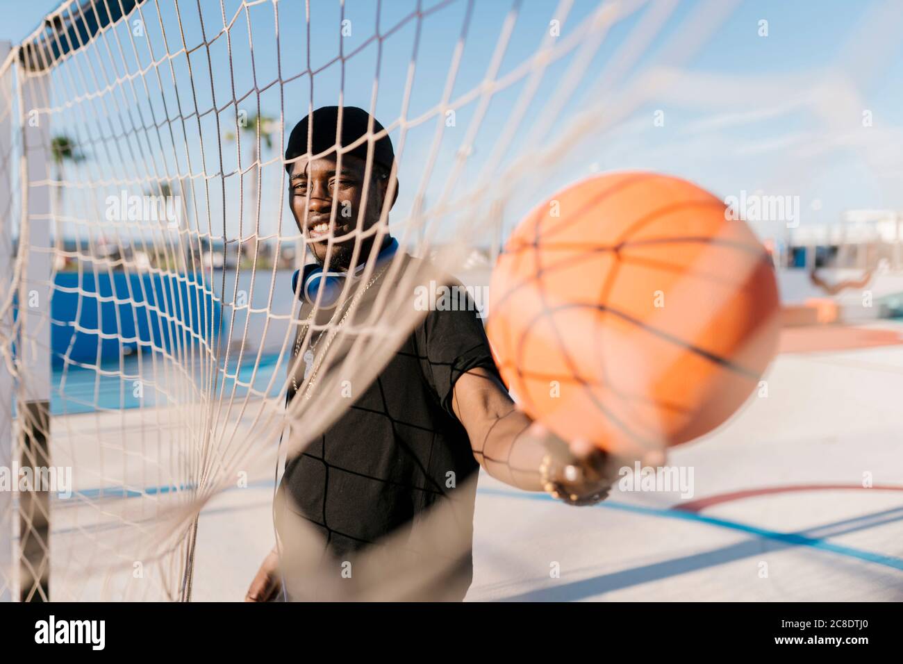 Junger Mann hält Basketball am Netz, während er vor Gericht steht An sonnigen Tagen Stockfoto