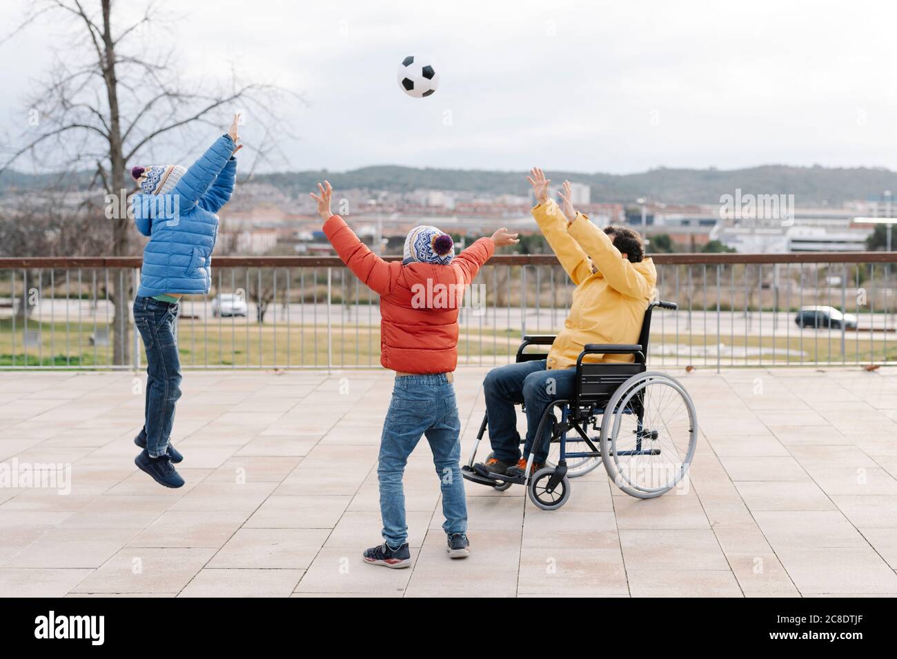 Spielerischer Vater sitzt auf dem Rollstuhl und wirft Ball auf begeisterte Söhne Im Park Stockfoto