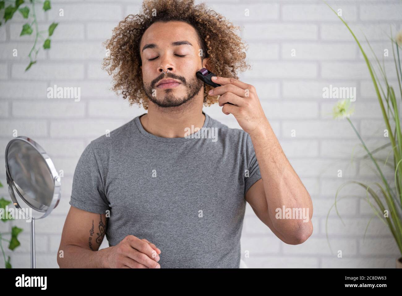 Junger Mann mit geschlossenen Augen massiert mit Mikroneedler im Gesicht Zu Hause Stockfoto