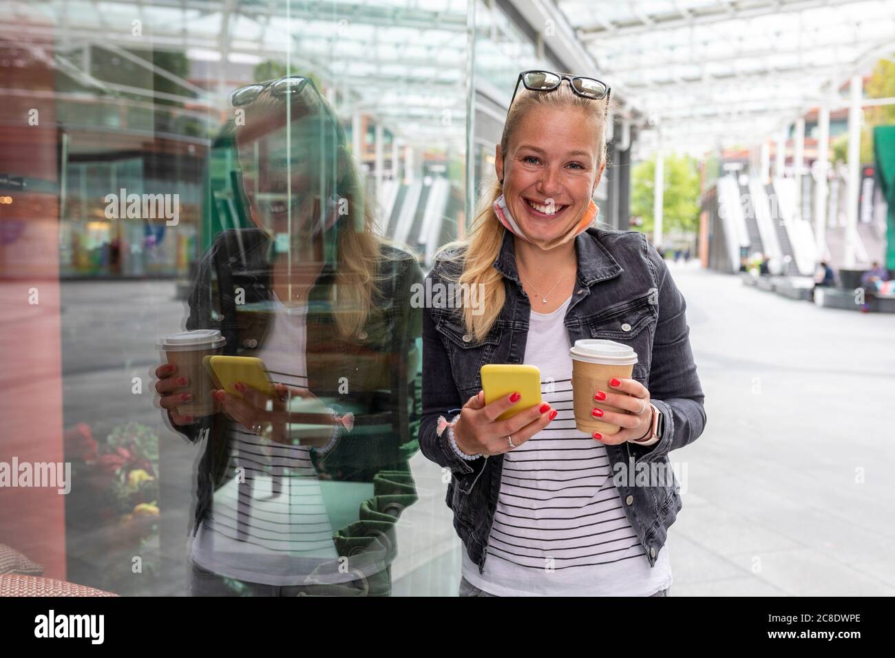 Lächelnde Frau hält Kaffeetasse mit Smartphone im Stehen Auf dem Bürgersteig in der Stadt Stockfoto