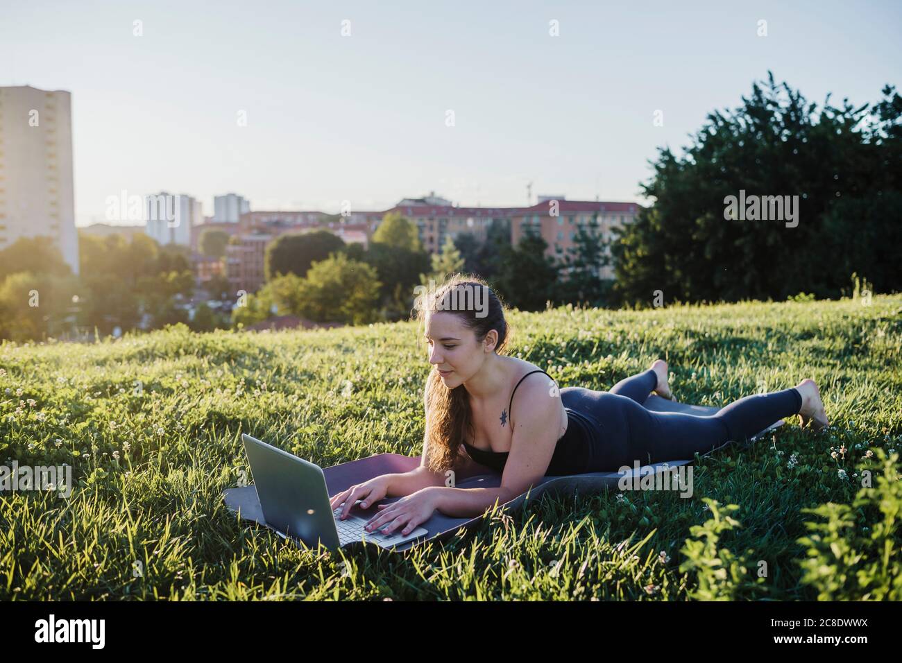 Sportliche junge Frau mit Laptop, während sie auf der Trainingsmatte liegt Im Stadtpark Stockfoto