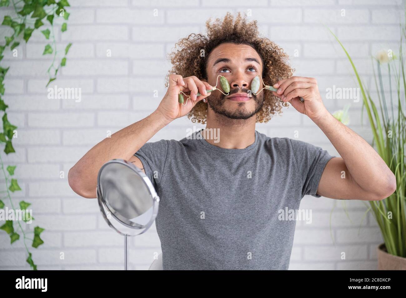 Junger Mann massiert Gesicht mit Jade Roller gegen Wand an Zu Hause Stockfoto
