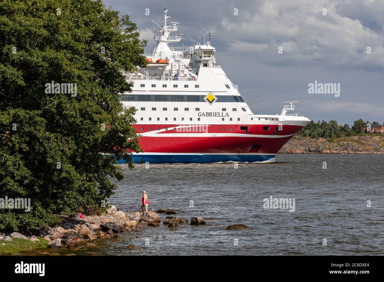 Frau in rosa Badeanzug kommt aus dem Wasser, während Kreuzfahrtschiff M/S Gabriella Lonna Insel in Helsinki Archipel, Finnland passiert Stockfoto