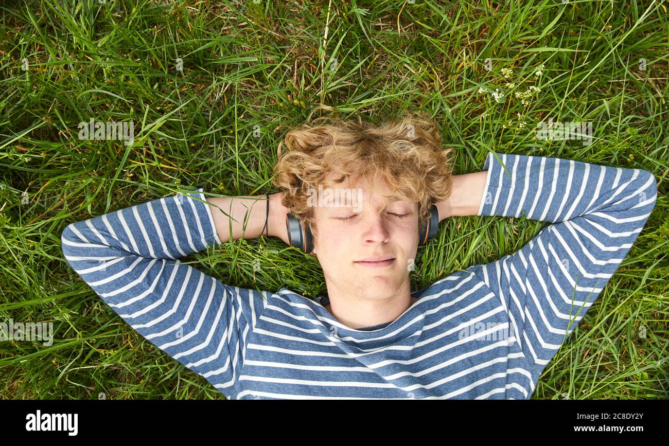 Porträt eines jungen Mannes mit lockigen blonden Haaren liegend Eine Wiese, die Musik mit Kopfhörern hört Stockfoto