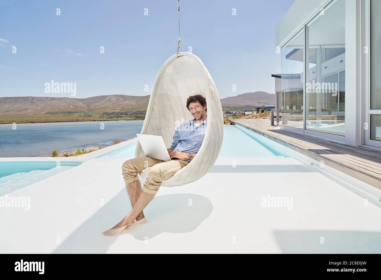 Mann, der mit einem Laptop in einem Hängesessel über dem Pool sitzt Stockfoto
