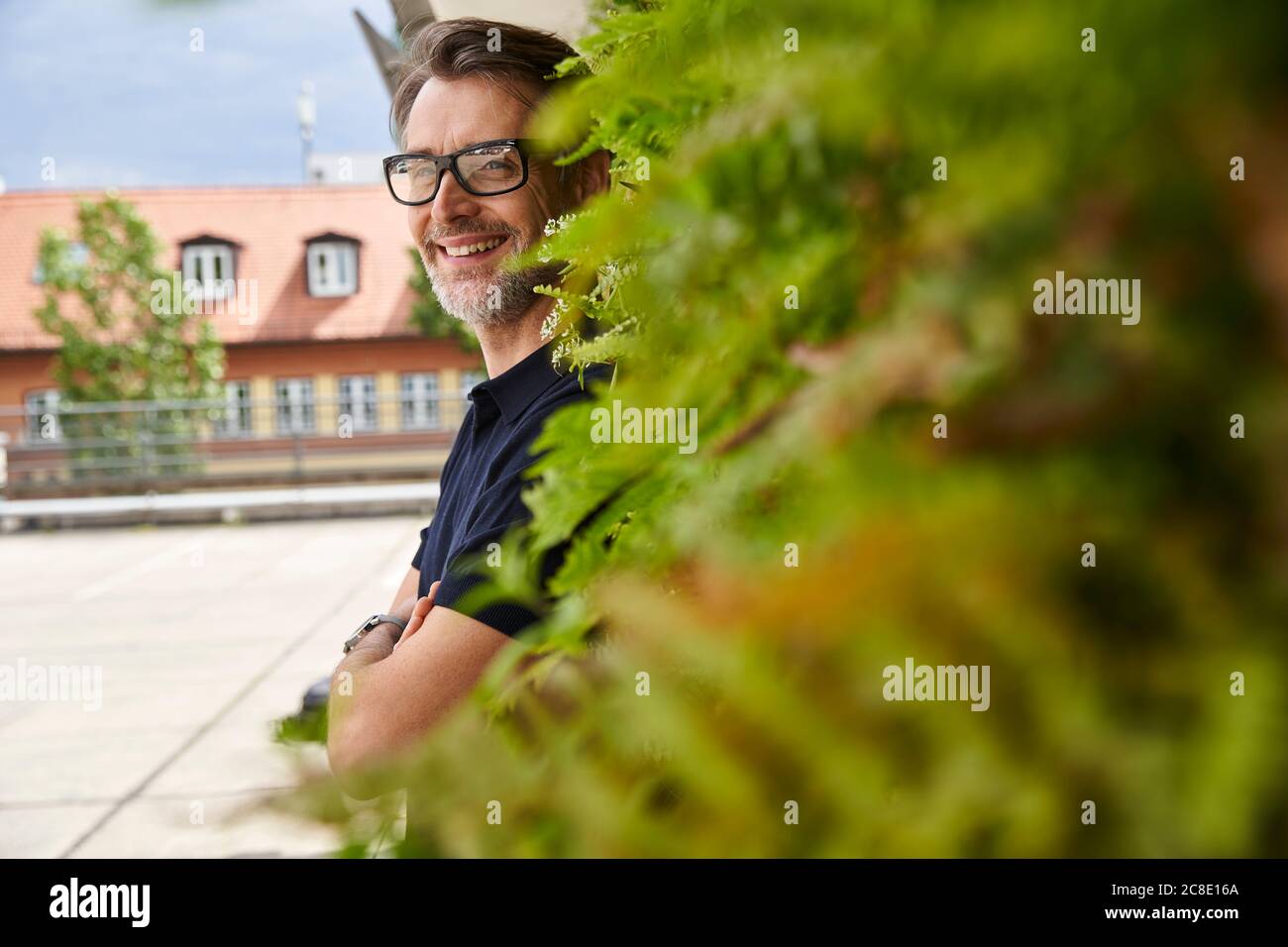 Nachdenklicher reifer Mann lächelt, während er bei Pflanzen auf dem Fußweg steht Am Hof Stockfoto