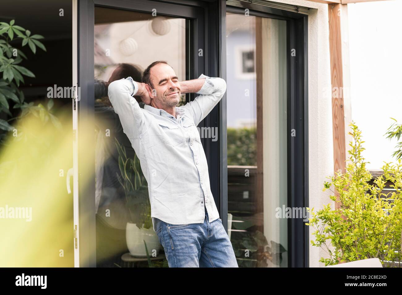 Entspannter reifer Mann mit Händen hinter dem Kopf auf Fenster gelehnt Im Hinterhof Stockfoto