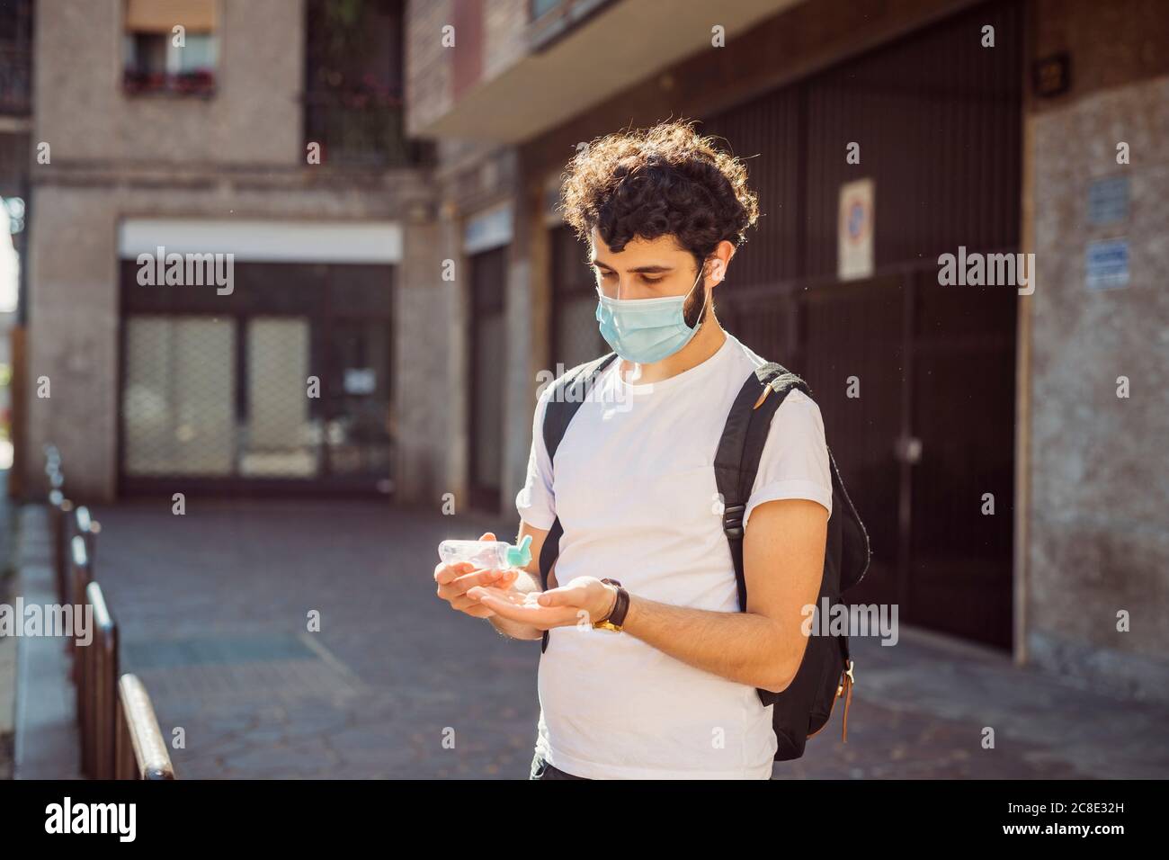 Junger Mann, der im Stehen die Hände mit Desinfektionsmittel wascht, trägt eine Maske Gegen Gebäude Stockfoto