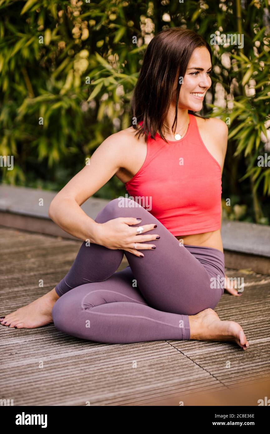 Lächelnde Frau beim Training, während sie auf Hartholzboden gegen Pflanzen sitzt Stockfoto