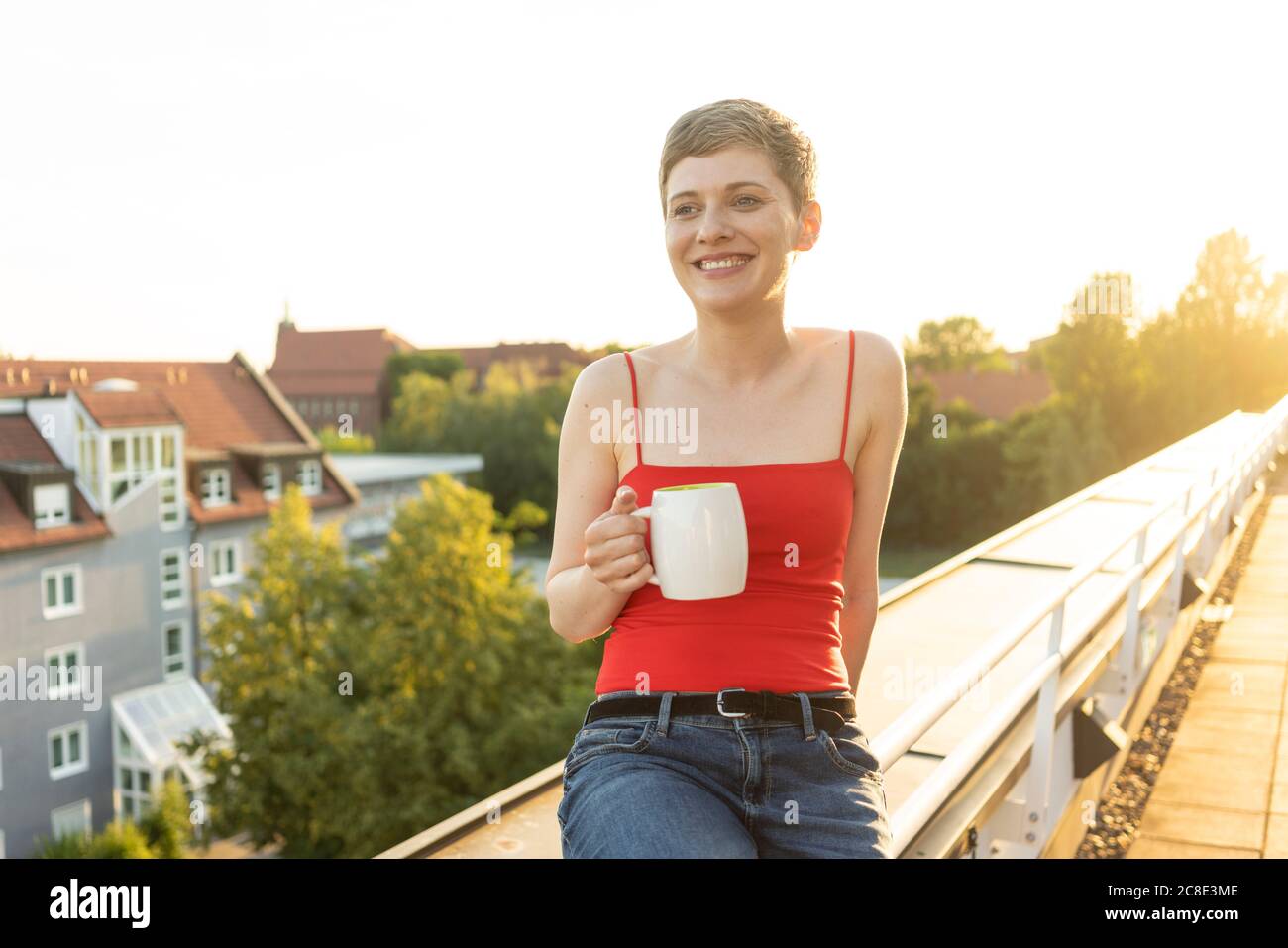 Lächelnde Frau mit kurzen Haaren hält Teebecher während des Sitzens Auf dem Geländer im Balkon bei Sonnenuntergang Stockfoto