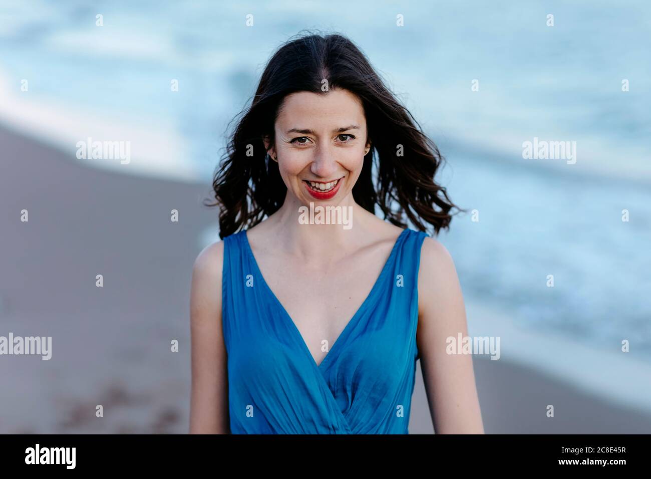 Dunkelhaarige Frau in blauem Kleid zu Fuß am Strand, lächelnd, Porträt Stockfoto
