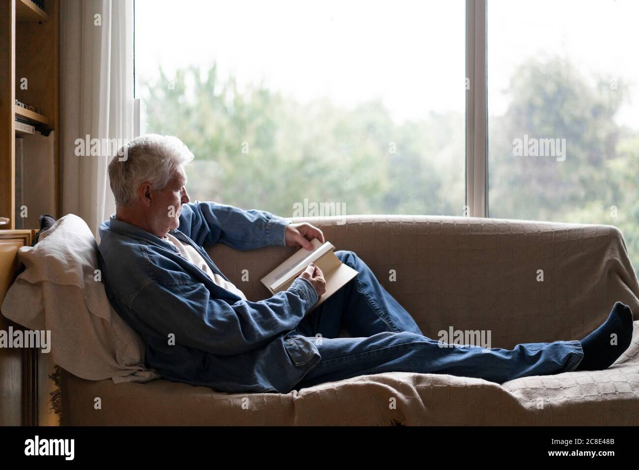 Ein älterer Mann liest ein Buch und entspannt sich auf dem Sofa am Fenster Zu Hause Stockfoto
