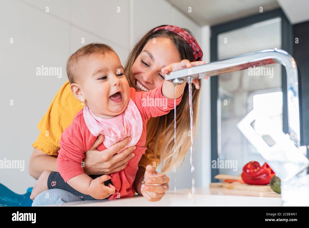 Close-up von lächelnden Mutter hält niedlichen Baby Mädchen spielen mit Wasserhahn in der Küche Stockfoto