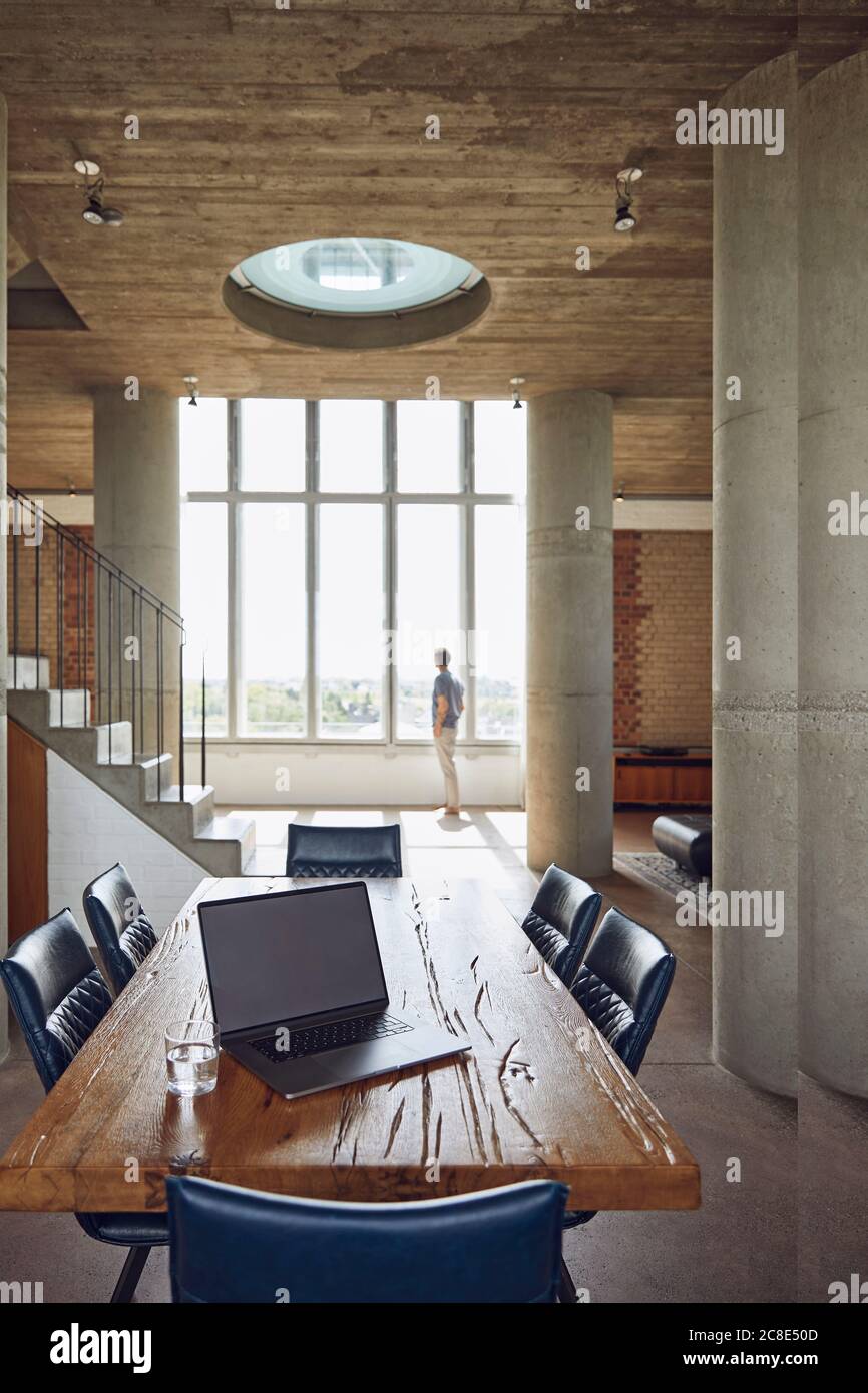 Laptop auf Holztisch in einer Loft-Wohnung mit Mann Am Fenster im Hintergrund Stockfoto