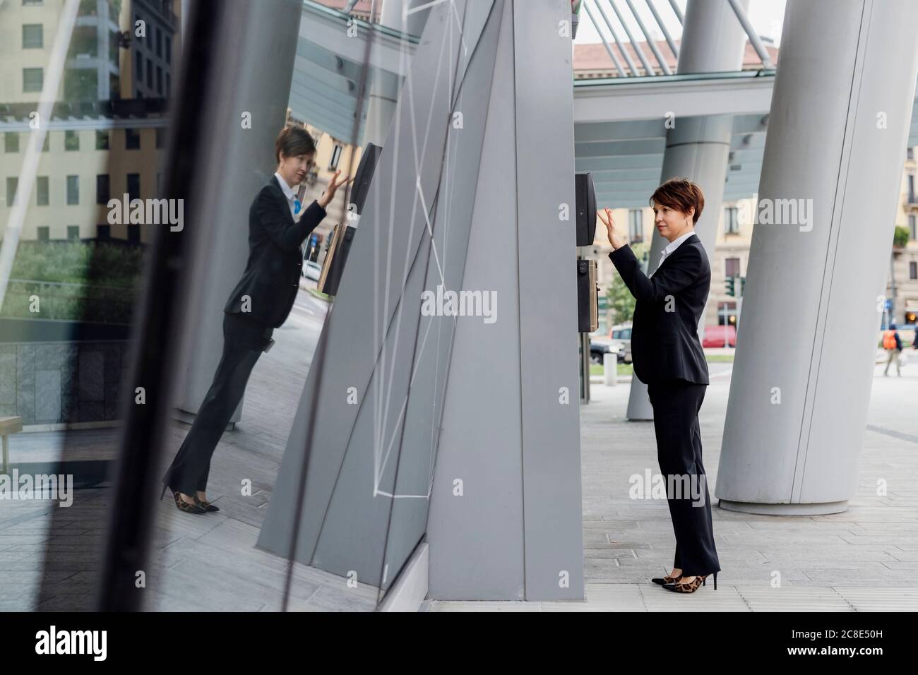 Geschäftsfrau mit kurzen Haaren mit ATM während Reflexion auf moderne Gebäude in der Stadt Stockfoto
