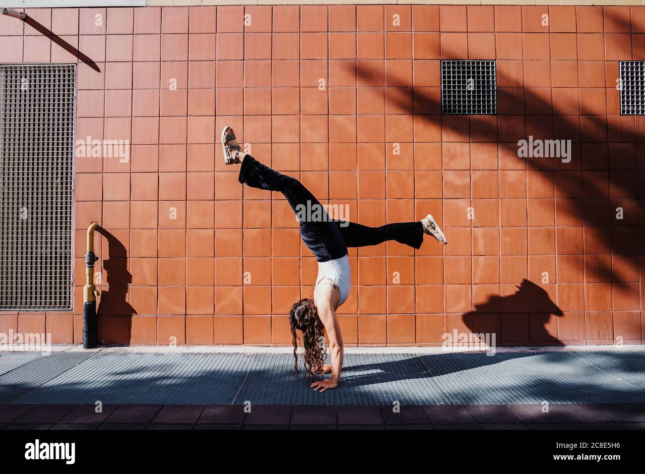 Junge Frau, die Handstand auf dem Bürgersteig gegen geflieste Wand in Stadt an sonnigen Tagen Stockfoto
