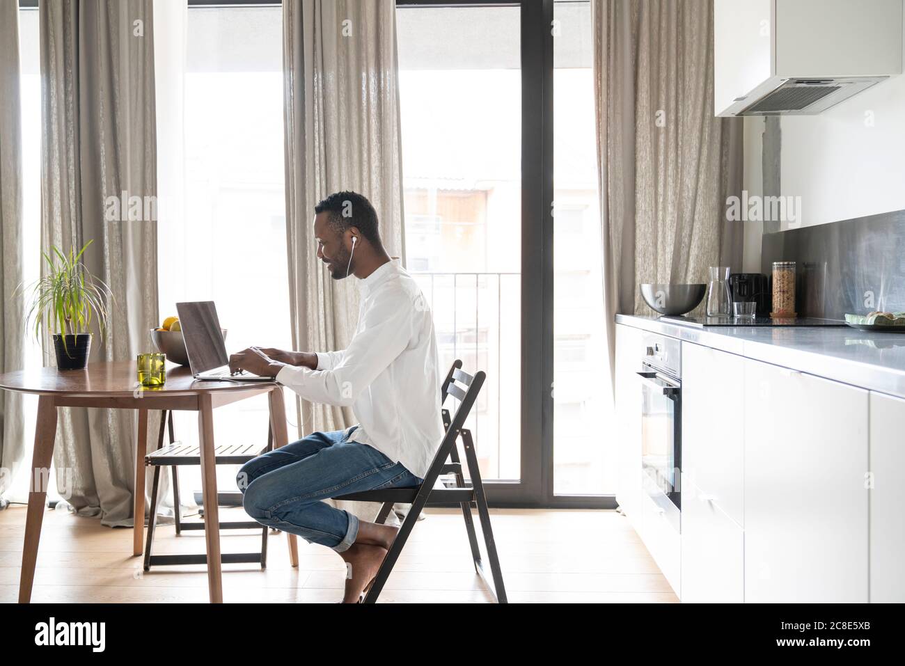 Lächelnder Mann, der in einem modernen Apartment mit einem Laptop am Tisch sitzt Und Ohrhörer Stockfoto