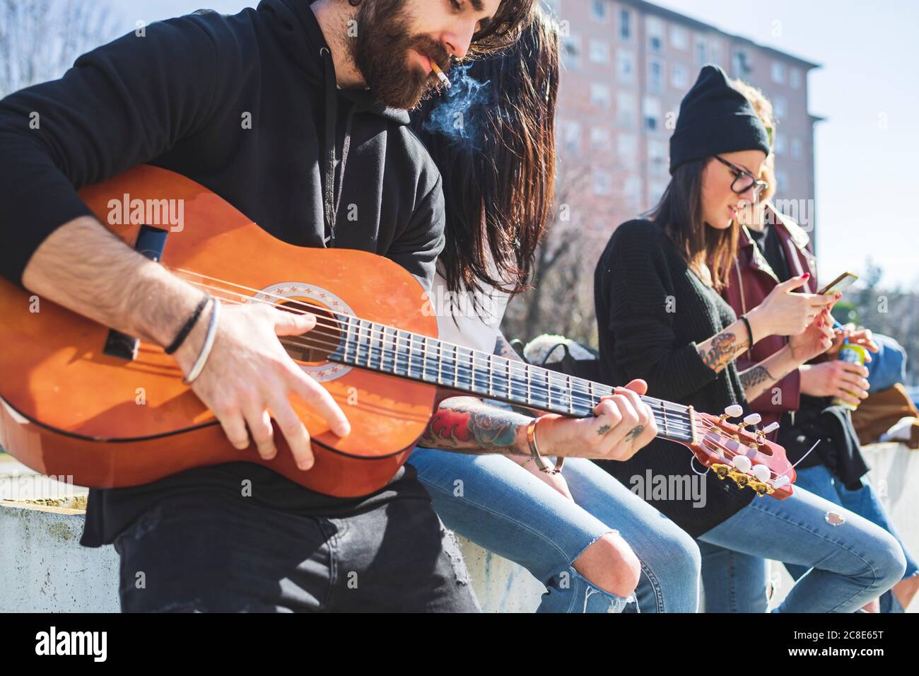 Mann, der Zigarette raucht und Gitarre spielt, während er mit Freunden chillte Stadt Stockfoto