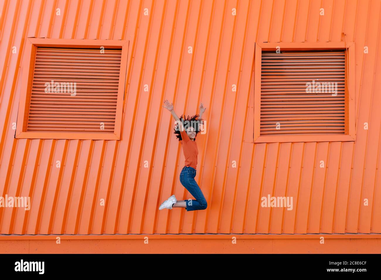 Fröhliche junge Frau, die mit den Armen gegen die orange Wand springen Stockfoto