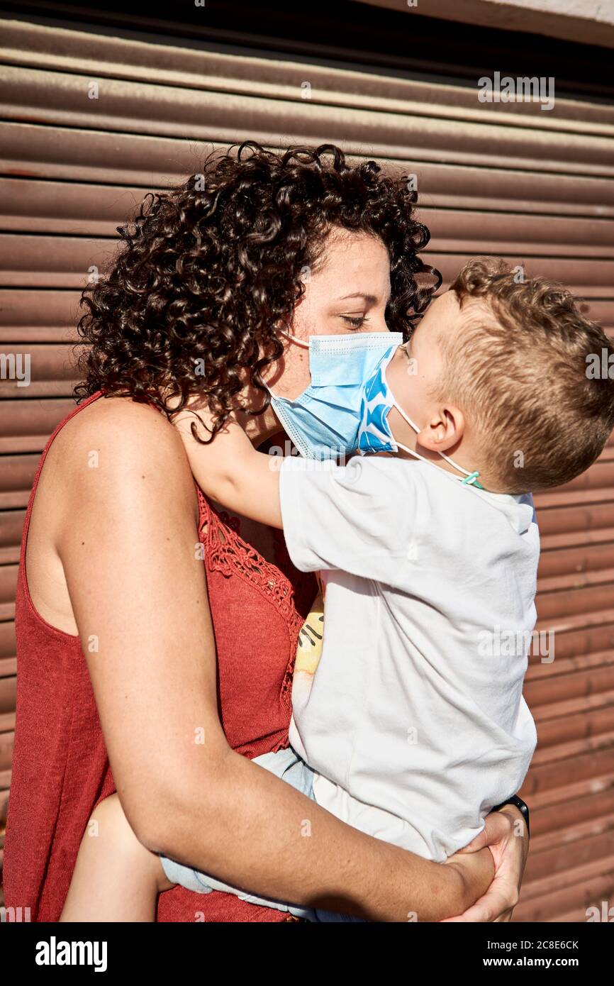 Mutter trägt Sohn mit Masken küssen ihn, während stehen gegen Geschlossener Verschluss Stockfoto