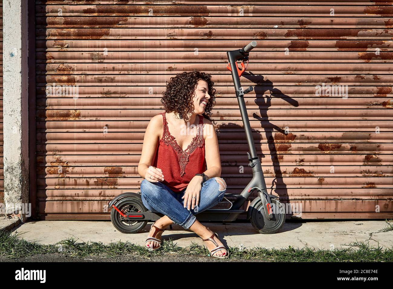 Lächelnde Frau mit lockigen Haaren sitzt auf Elektro-Push-Roller Gegen geschlossenen Verschluss Stockfoto