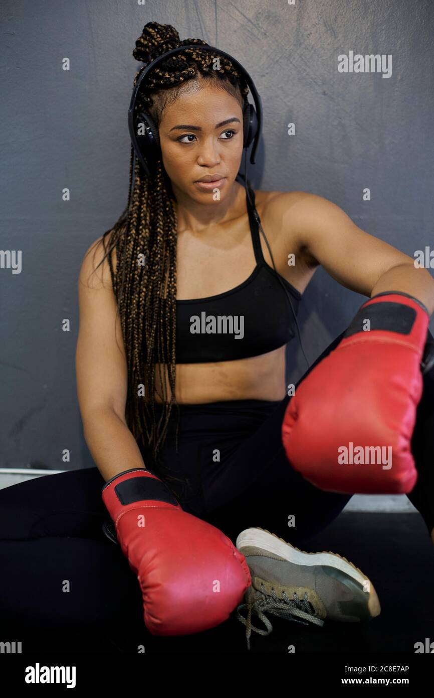 Müde Sportlerin trägt rote Handschuhe und hört Musik, während sie sitzt Gegen die Wand im Fitnessraum Stockfoto