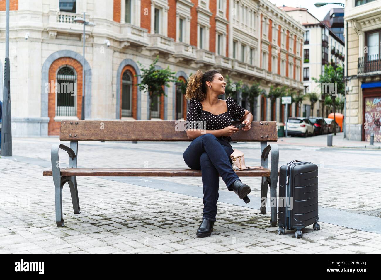 Lächelnd junge Geschäftsfrau in smart casual sitzen auf Bank by Gepäck Stockfoto
