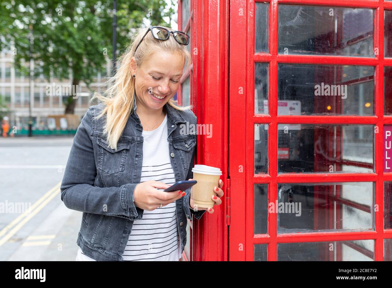 Lächelnde Frau hält Kaffeetasse mit Smartphone im Stehen Mit Telefonzelle Stockfoto