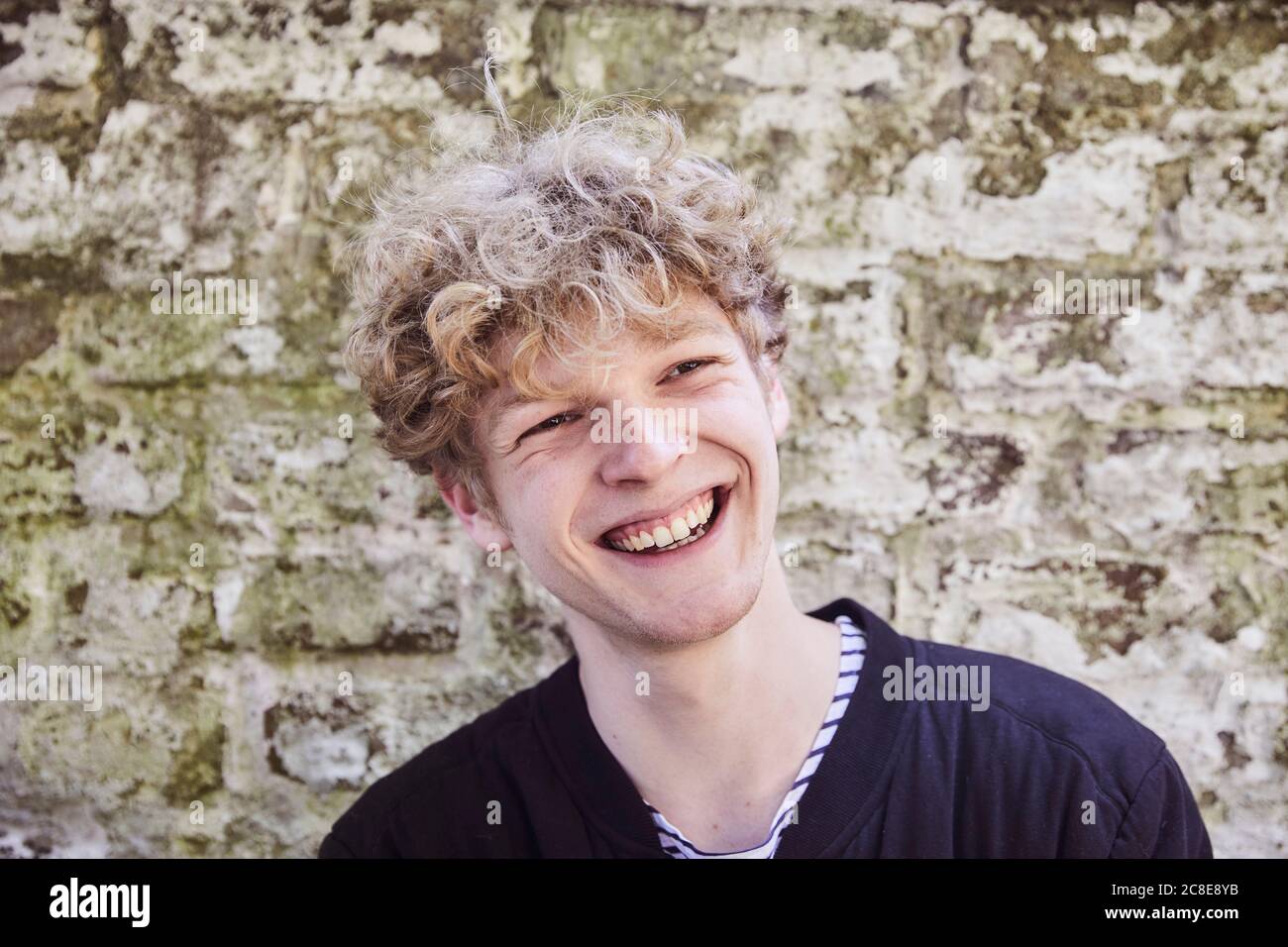 Porträt des lachenden jungen Mannes mit lockigen blonden Haaren in Vorderseite der verwitterten Wand Stockfoto