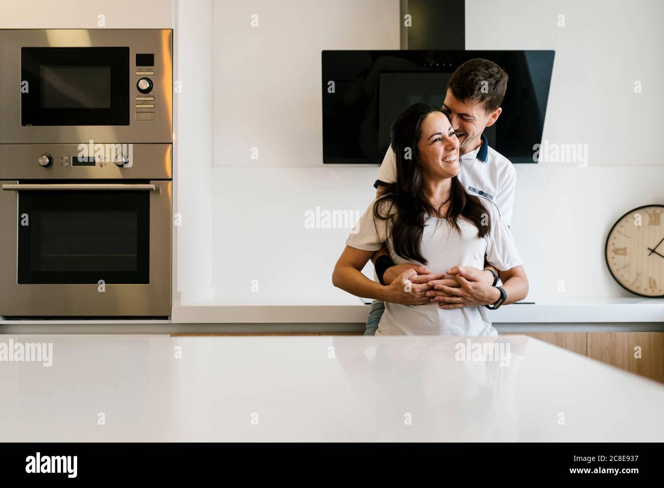 Lächelnder Mann umarmt Frau von hinten in Küche Stockfoto