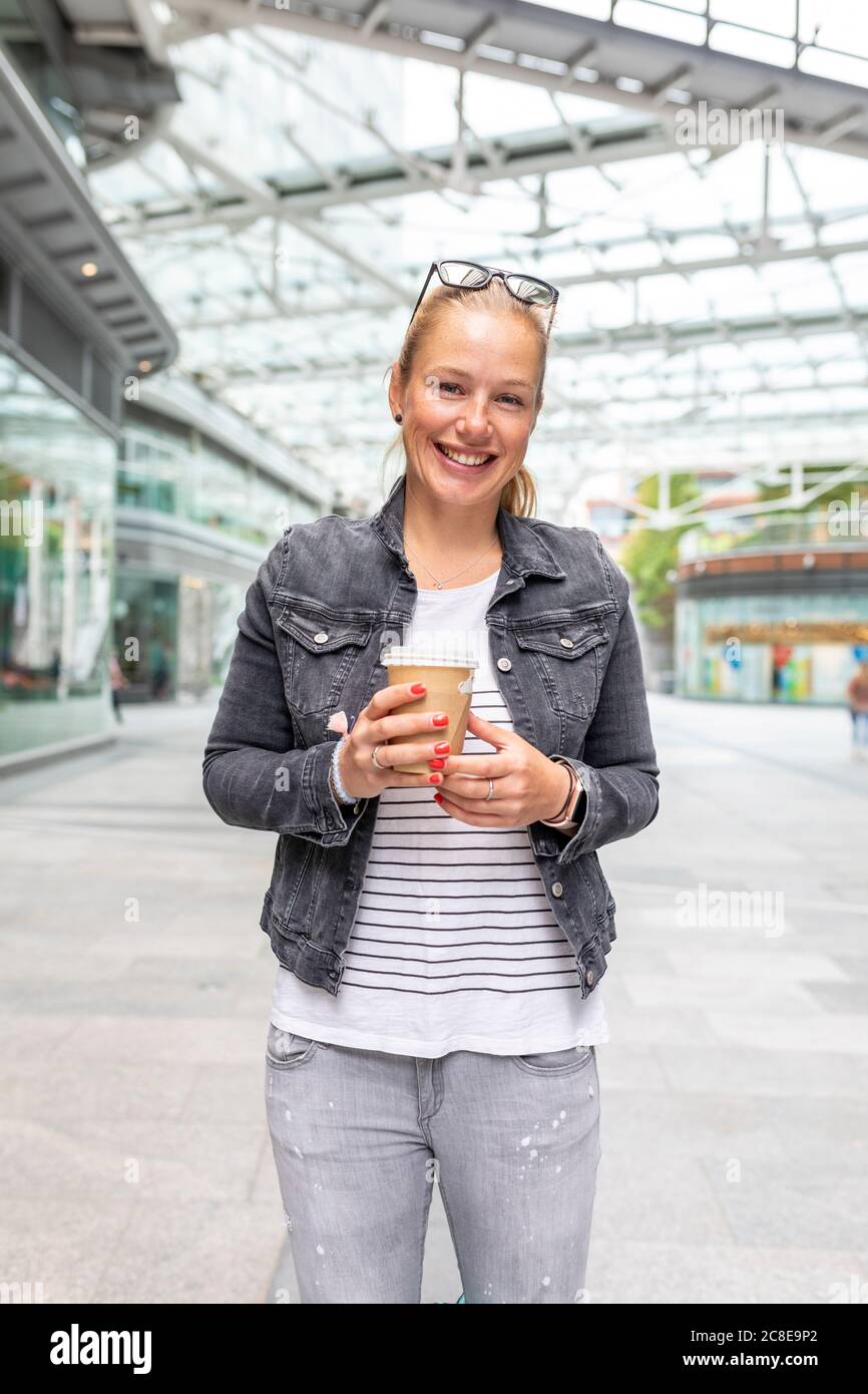 Lächelnde Frau mittleren Erwachsenen hält Kaffeetasse, während Sie auf stehen Bürgersteig in der Stadt Stockfoto
