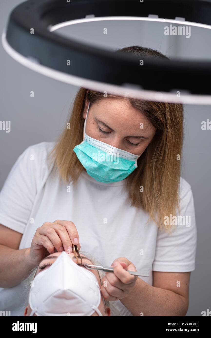 Kosmetikerin trägt Gesichtsmaske, während mit Werkzeugen auf Kunden für Wimpernverlängerung im Beauty Spa Stockfoto