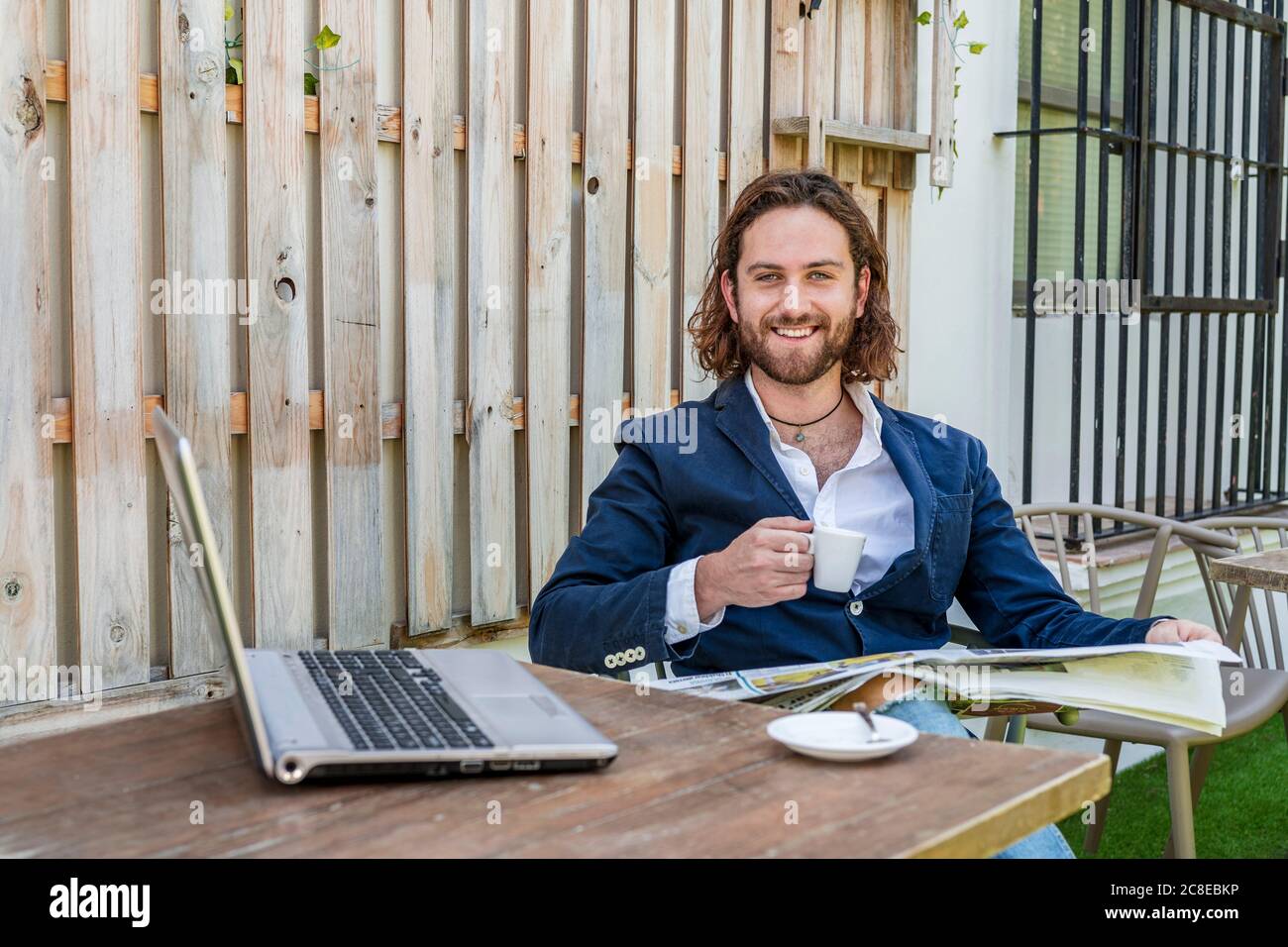 Lächelnd hübscher junger Geschäftsmann, der Zeitung liest, während er die Kaffeetasse hält Mit dem Laptop am Tisch im Café Stockfoto