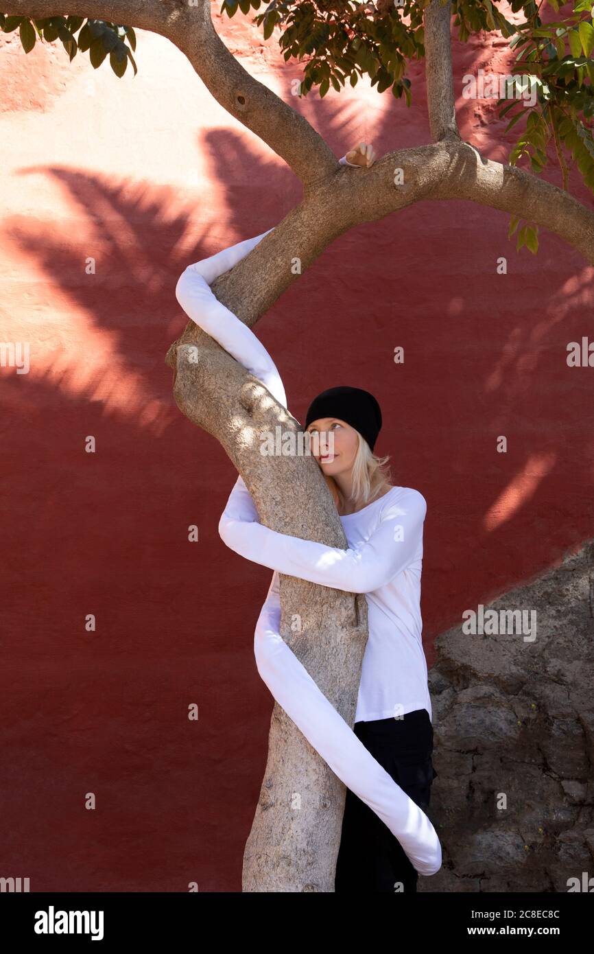 Nachdenkliche Frau mit künstlichen langen Händen umarmen Baumstamm gegen Wand Stockfoto