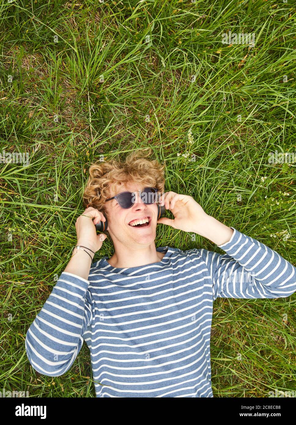 Porträt eines lachenden jungen Mannes mit lockigen blonden Haaren liegend Auf einer Wiese Musik hören mit Kopfhörern Stockfoto