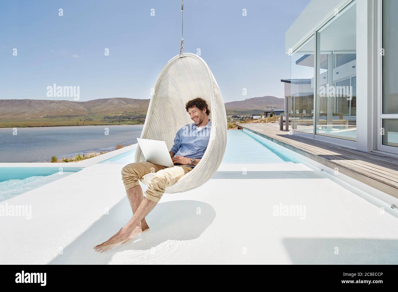 Mann, der mit einem Laptop in einem Hängesessel über dem Pool sitzt Stockfoto