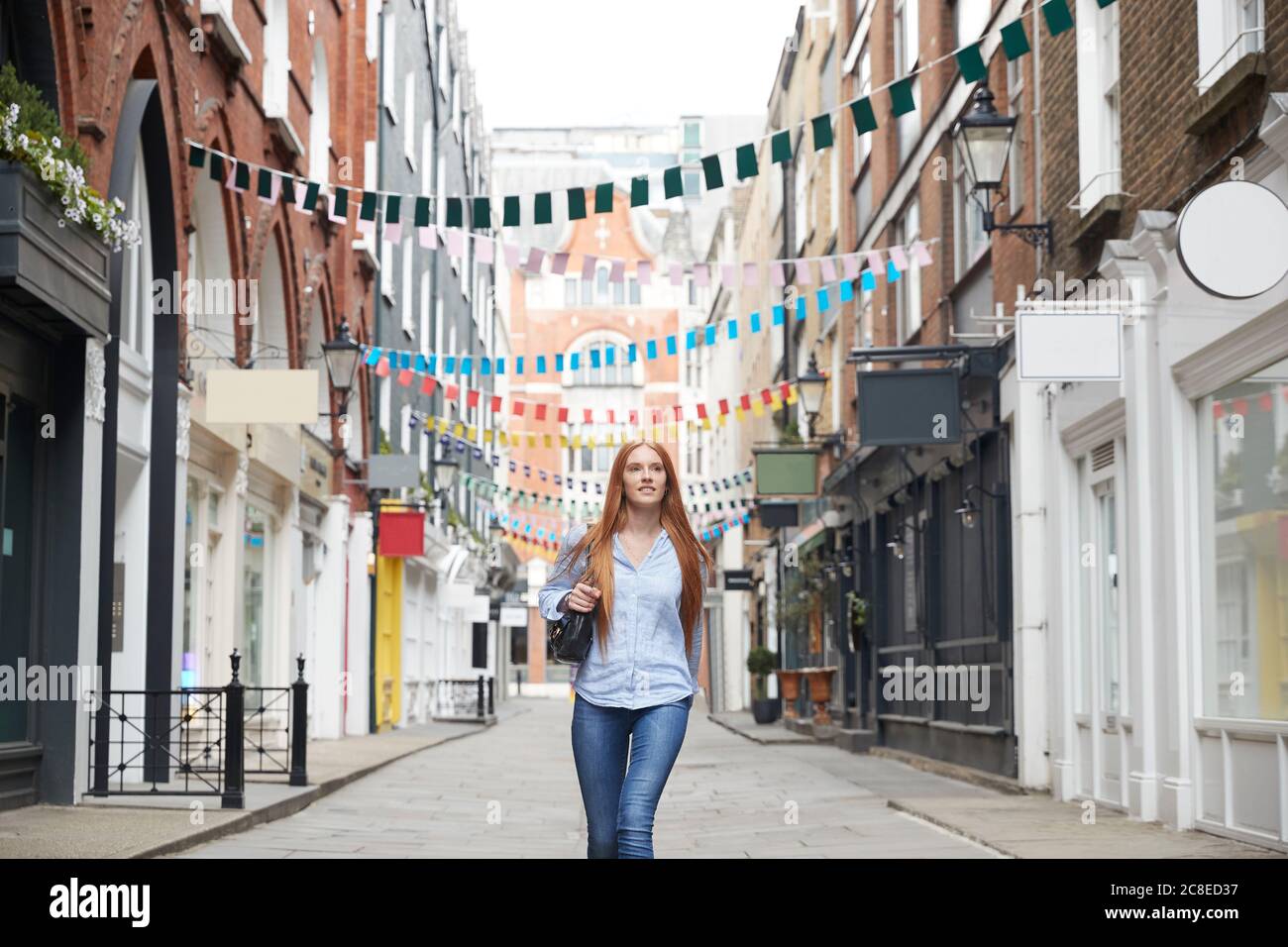 Schöne Rothaarige Frau, die auf einer Gasse gegen die Bäuste inmitten von Gebäuden läuft In der Stadt Stockfoto