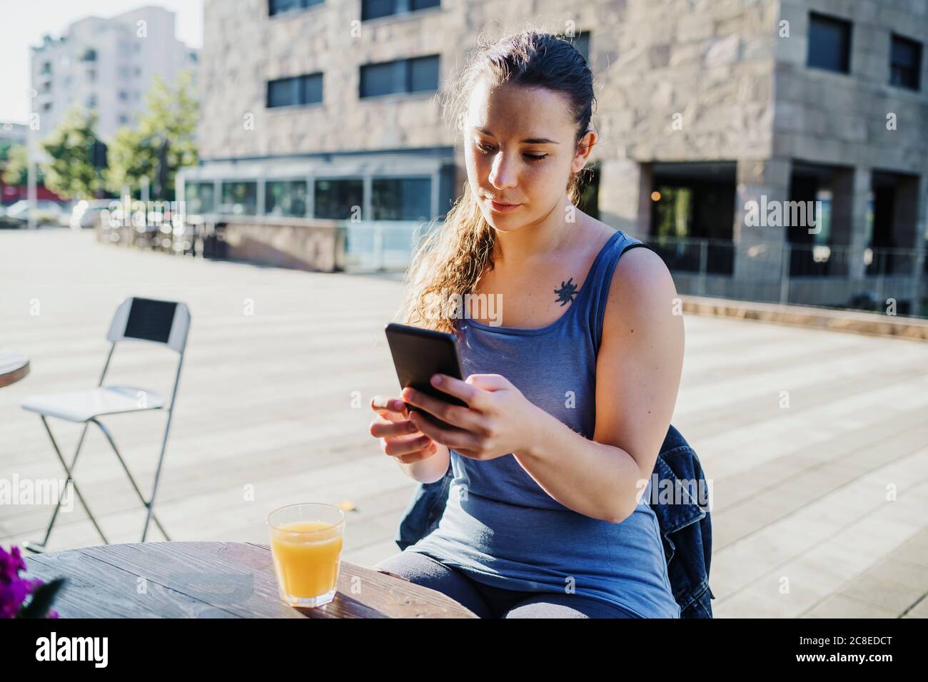 Sportliche junge Frau mit Smartphone, während sie auf dem Bürgersteig sitzt Cafe in der Stadt Stockfoto