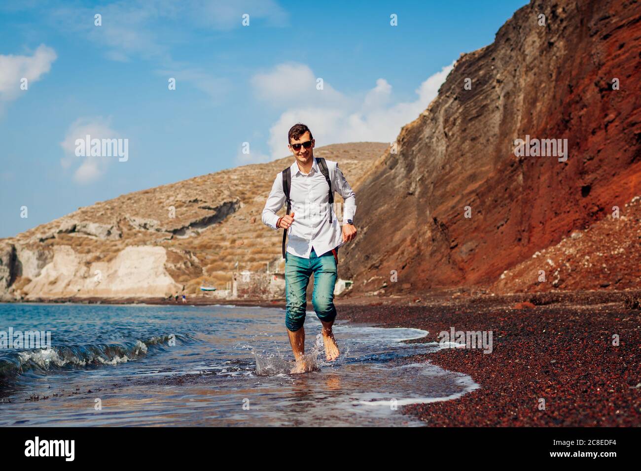 Fröhlicher Tourist, der barfuß am Roten Strand am Meer auf Santorini läuft. Mann mit Rucksack unterwegs Stockfoto