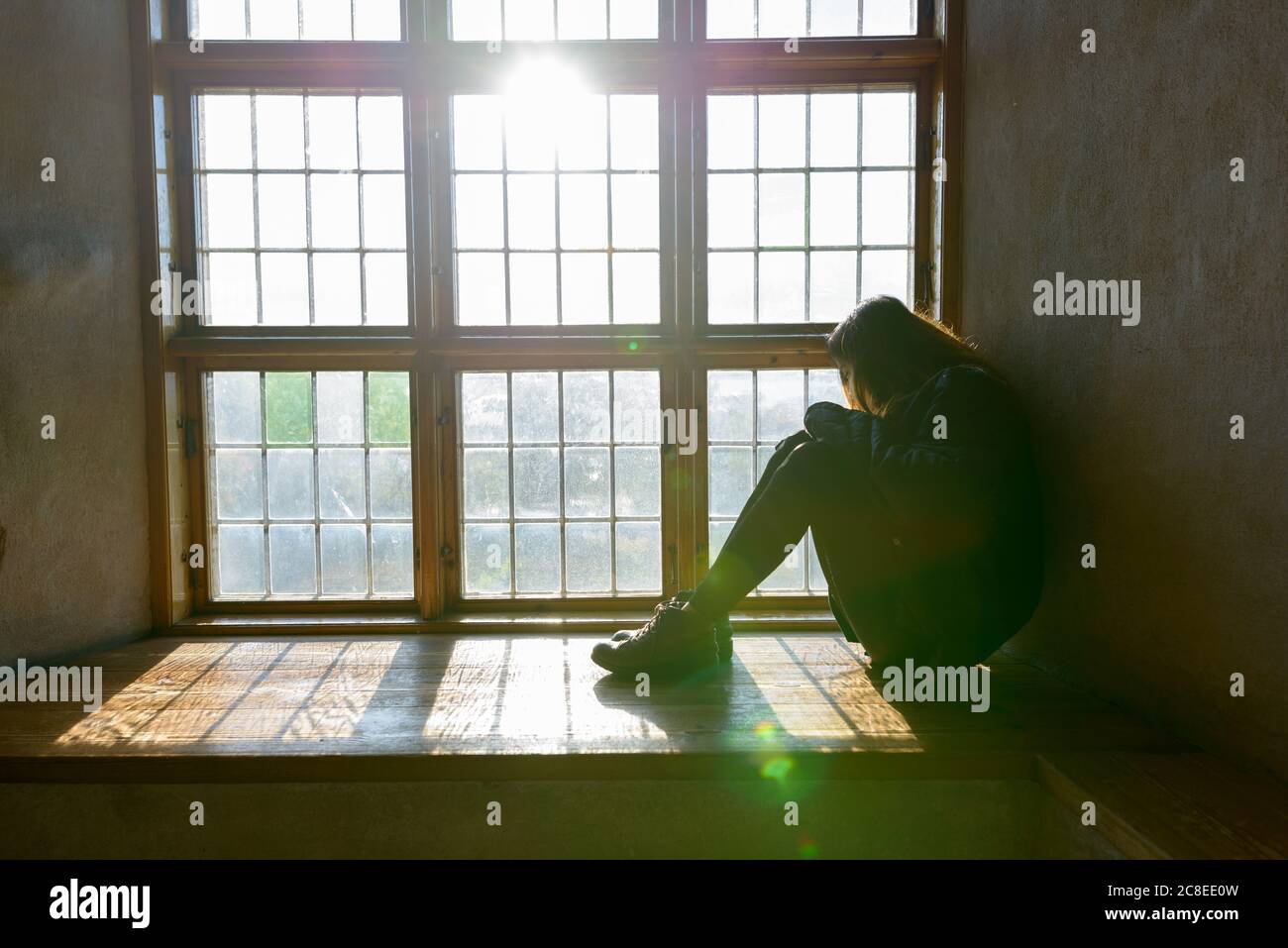 Junge Frau, die vor einem geschlossenen Holzfenster sitzt und das Sonnenlicht einströmt Stockfoto